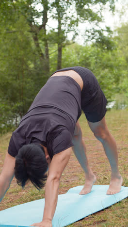 Video-Vertical-De-Un-Hombre-Vestido-Con-Ropa-Deportiva-Haciendo-Estiramientos-De-Yoga-Sobre-Una-Colchoneta-En-Un-Bosque-Junto-A-Un-Lago-O-Río-Disfrutando-De-La-Paz-Y-La-Belleza-De-La-Naturaleza-Filmado-En-Tiempo-Real-1